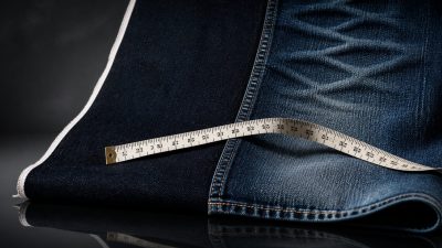 Stack of raw indigo jeans with one pair on a wooden hanger, tailor's measuring tape draped across, soft natural light on a da