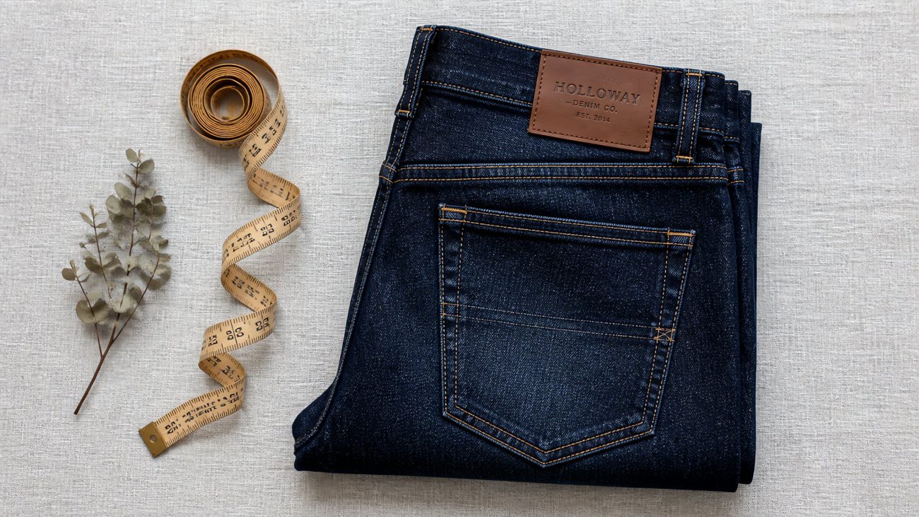 A neatly folded stack of indigo jeans with a tailor's measuring tape resting on top in soft natural light