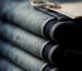 Stack of indigo jeans on a wooden shelf beside a tailor's measuring tape in soft natural light
