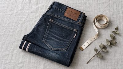 Stack of indigo jeans beside tailor tape on light wood, soft shadows, muted minimalist scene