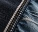 Folded stack of raw indigo jeans beside a washed pair on a wooden hanger with a tailor tape in soft natural light